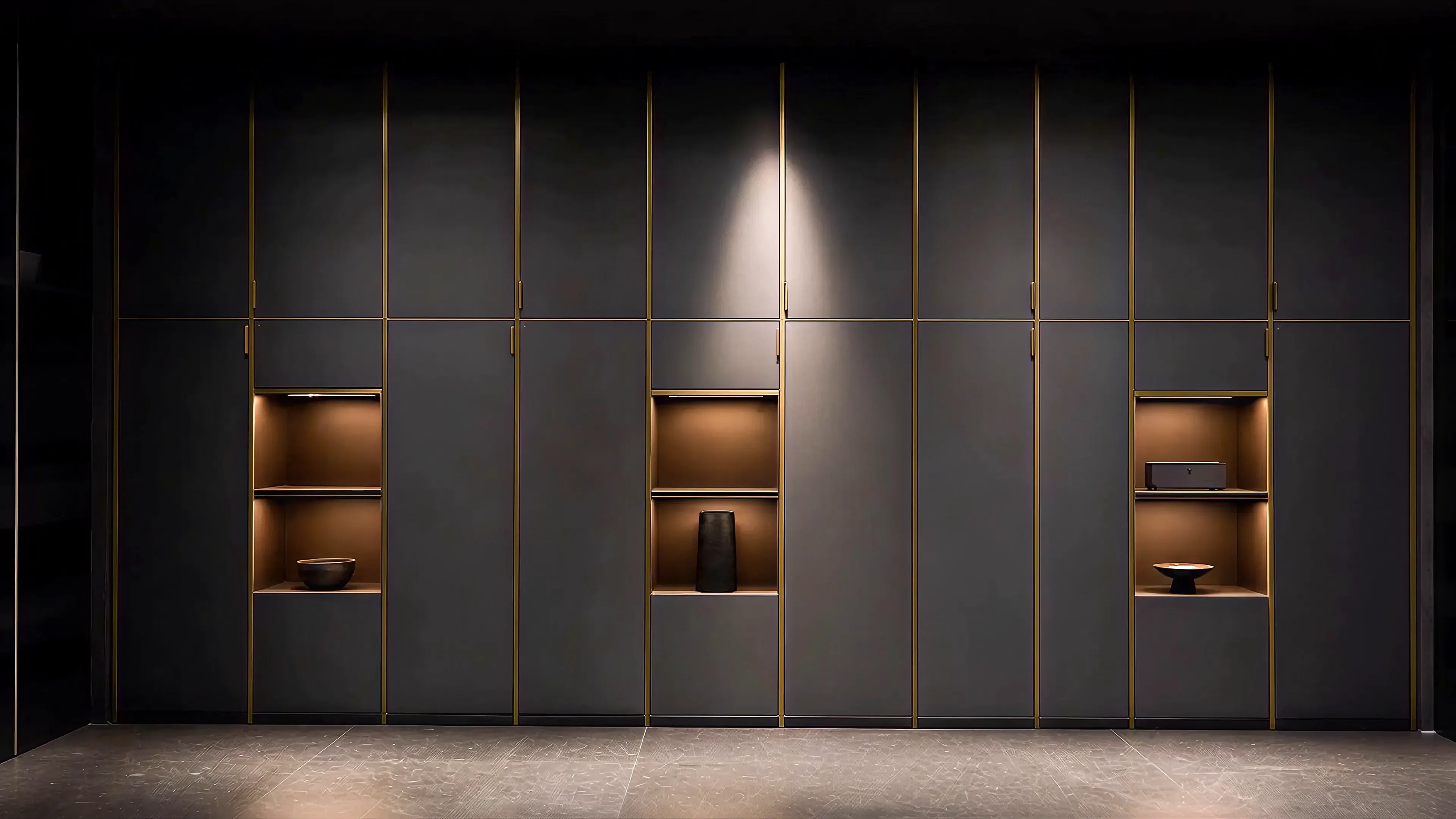 A fitted storage wall across a dark stage, charcoal fronts on a brass grid, three bays lit from inside, under one hard spotlight