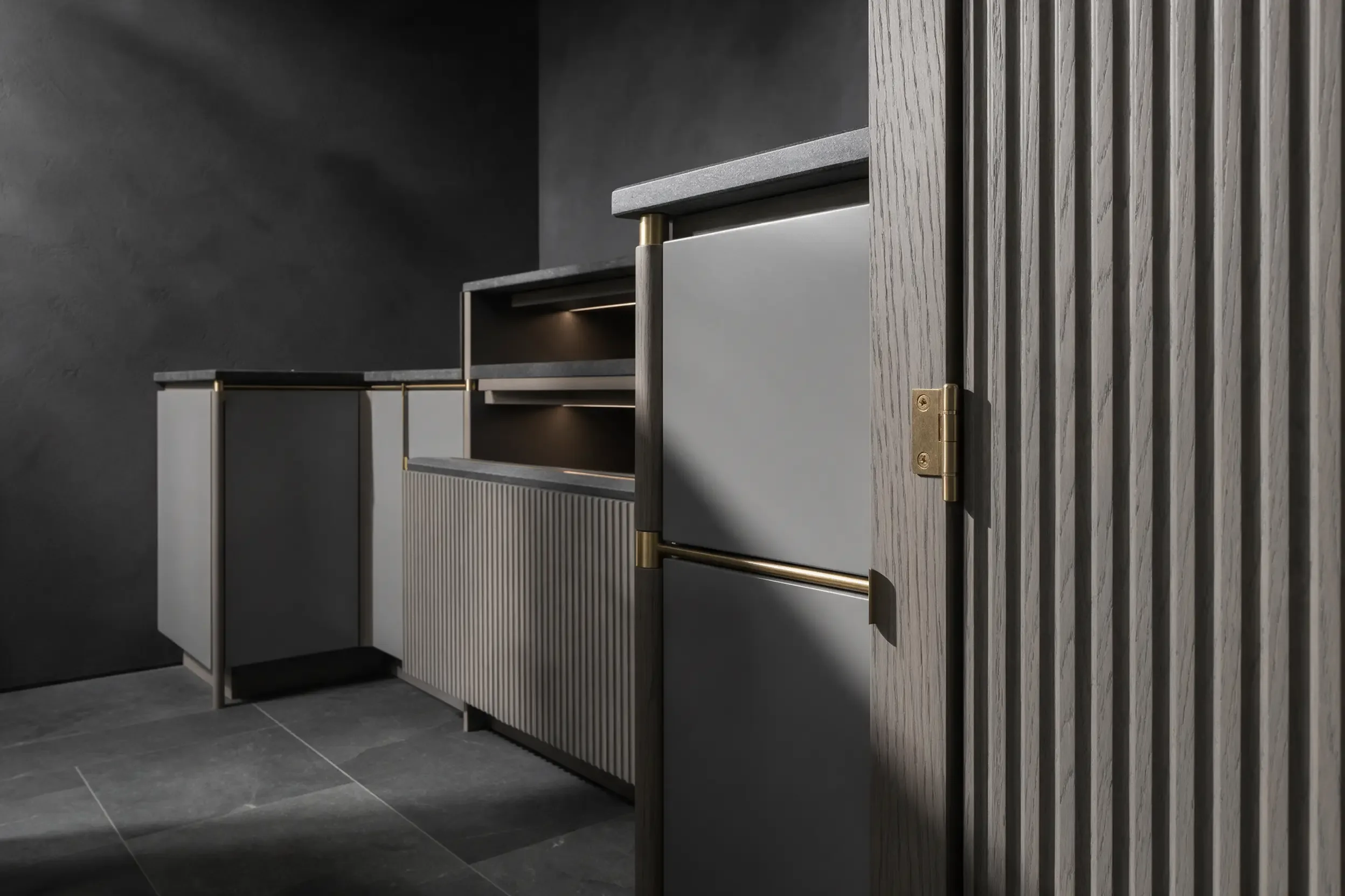 The corner of a fitted grey cabinet with a slim brass handle and a lit open bay