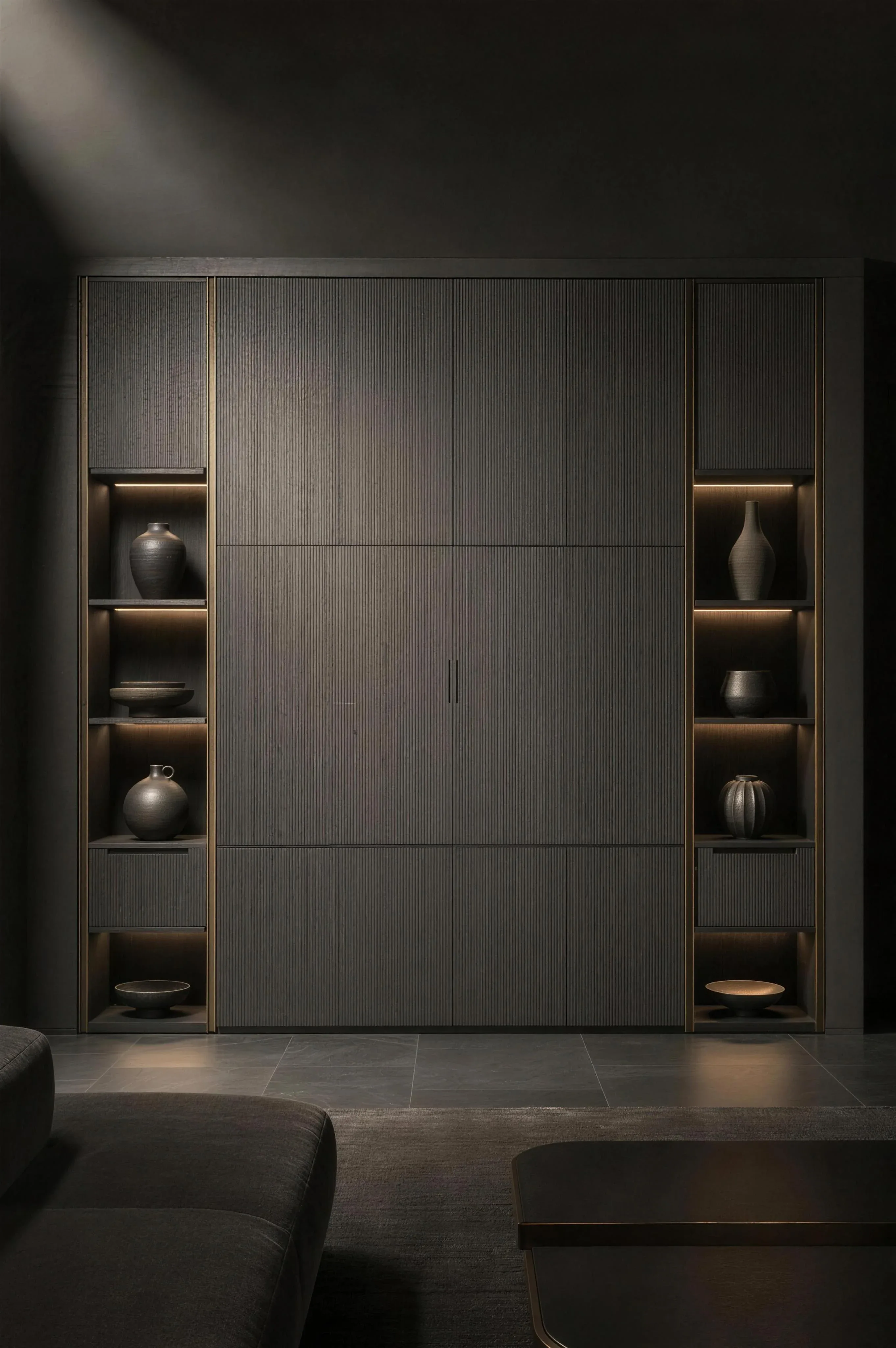 A floor to ceiling fitted storage wall in fluted grey with lit niches holding dark ceramics and brass rails, under one hard spotlight