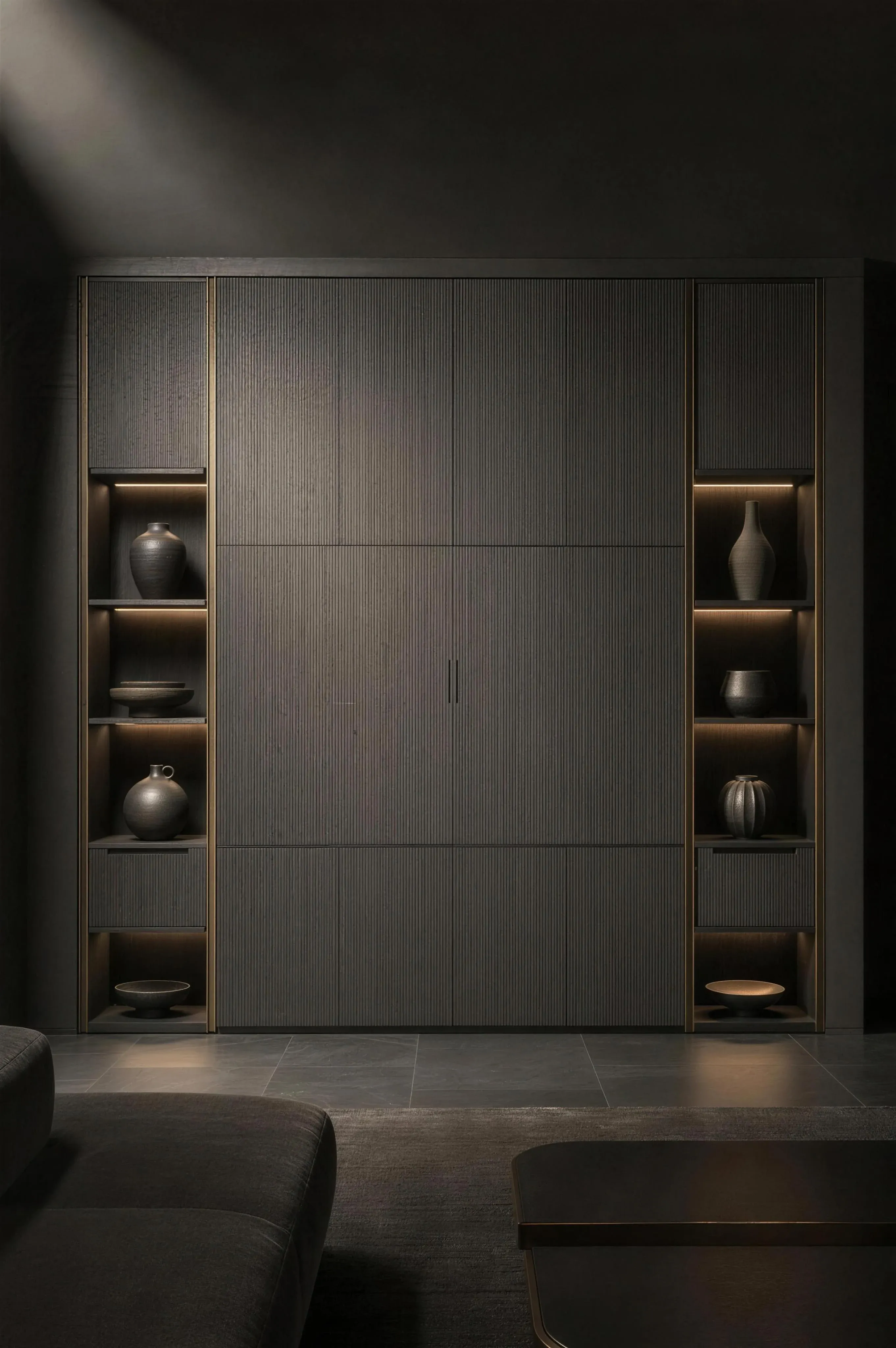 A floor to ceiling fitted storage wall in fluted grey with lit niches holding dark ceramics and brass rails, under one hard spotlight