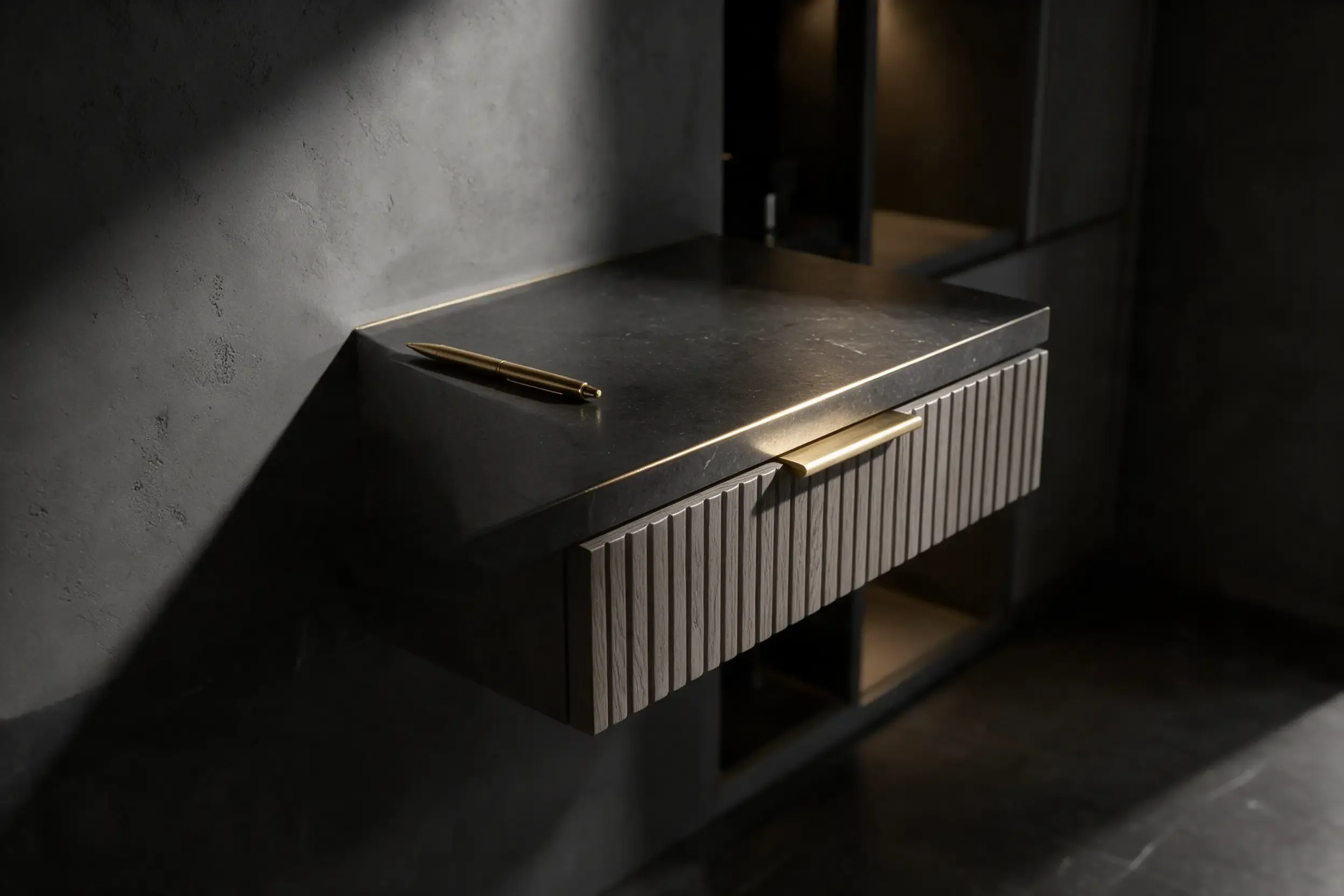 A dark stone desk top with a brass cable grommet and a fluted grey panel behind, in raking light