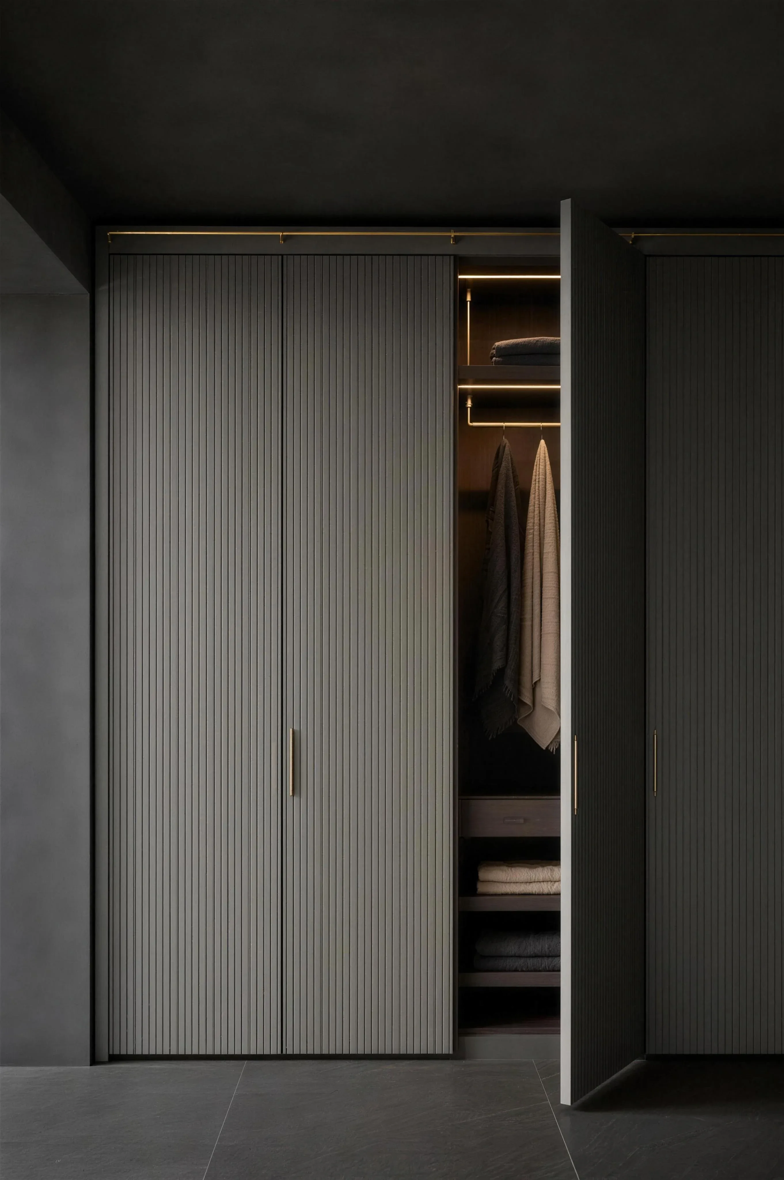 A floor to ceiling fitted wardrobe in fluted grey with brass rails, one tall door open on lit shelves and hanging linen, under one hard spotlight